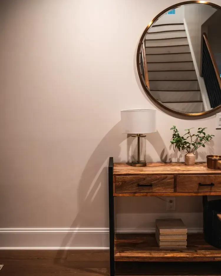 Wall mirror above a rustic console table with lamp, plant, and books in a neutral hallway