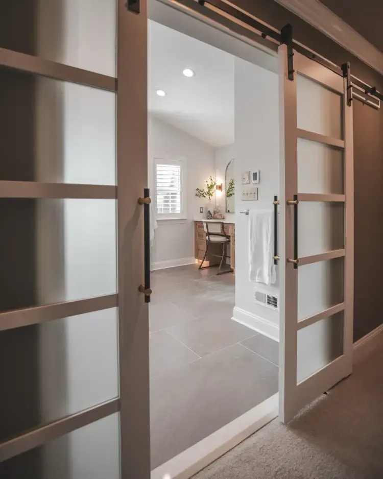 Open barn doors reveal a bright bathroom with white walls, gray floor, and towel hooks.
