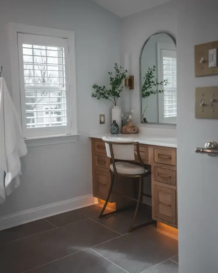 Bright bathroom vanity with mirror, chair, plant, and window with white blinds and gray walls