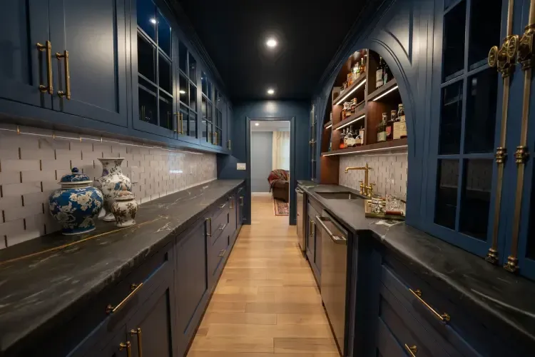 Narrow navy kitchen with gold hardware, long counters, wood floor, and built-in shelves lit by ceiling lights