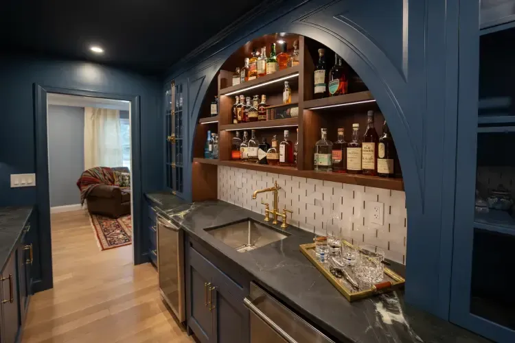 Blue arched bar nook with shelves of liquor and a sink, opening to a hallway beyond