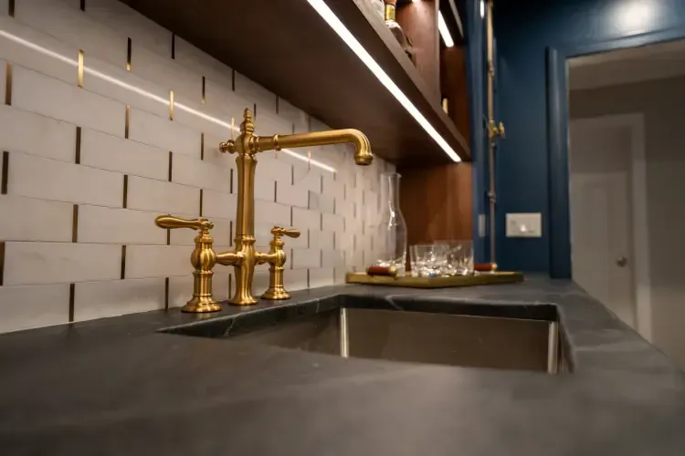 Modern kitchen sink with gold faucet, white tiled backsplash, and dark countertop near a doorway