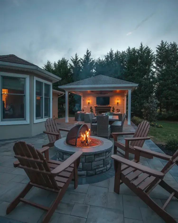 Backyard patio with a round fire pit, Adirondack chairs, and a lit outdoor pavilion at dusk