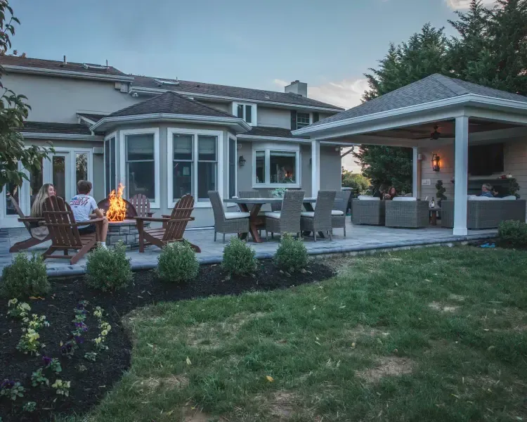 Backyard patio with fire pit, lounge chairs, and covered seating beside a house at dusk