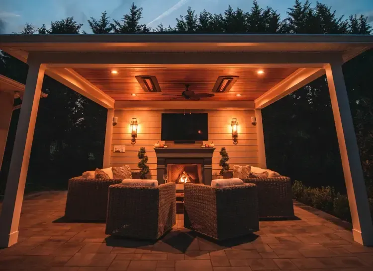 Cozy outdoor patio lounge with wicker seating, a fire pit, and warm overhead lighting at dusk