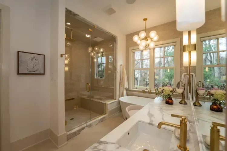 Bright bathroom with marble vanity, soaking tub, glass shower, brass fixtures, and a window view of trees.