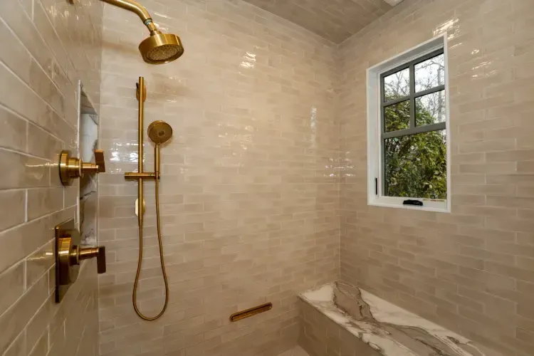 Accessible beige-tiled shower with gold fixtures, bench, and a window overlooking greenery