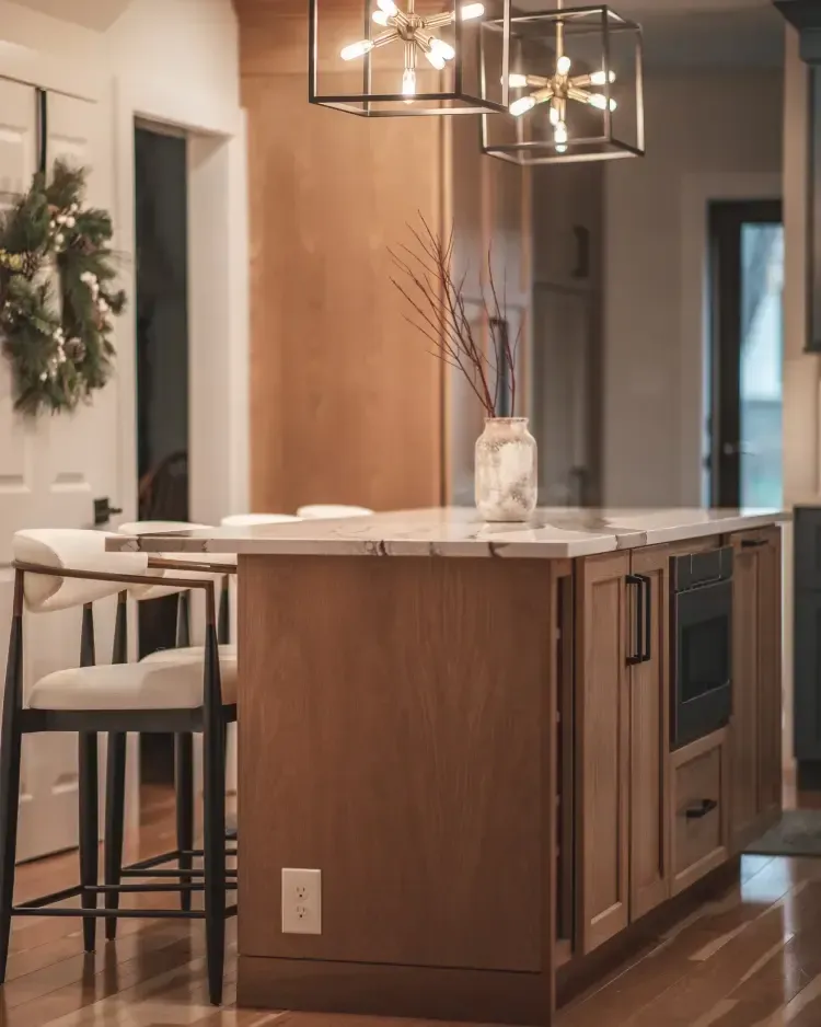 Modern kitchen island with wood cabinets, white countertop, pendant lights, and bar stools in a bright hallway.