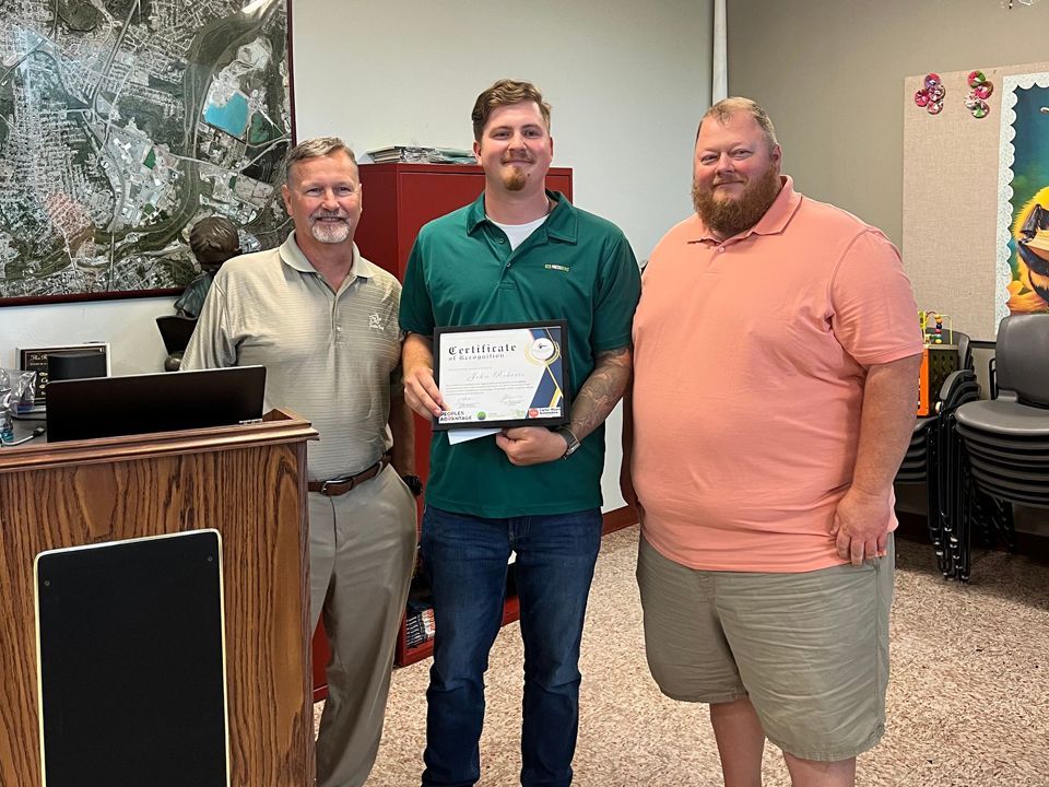 Three men pose indoors. A man in a green shirt holds a certificate, flanked by two others in tan and peach shirts. A map hangs in the background.