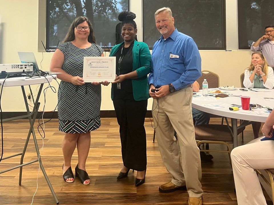 Three people stand indoors: a woman in a black and white dress holds a certificate, a woman in a green jacket, and a man in a blue shirt smiles.