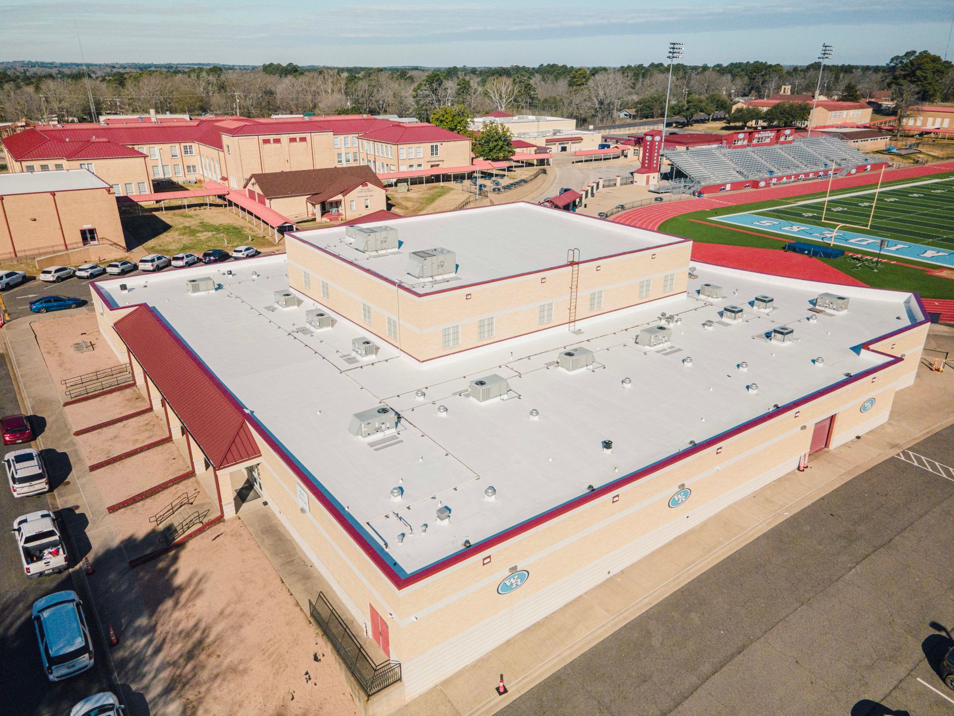 An aerial view of a large building with a white roof and a football field in the background.