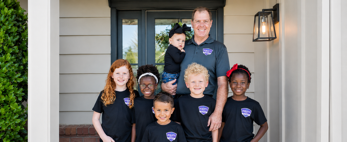 Family of six posing on a front porch, smiling in matching dark shirts beside a man holding a child.