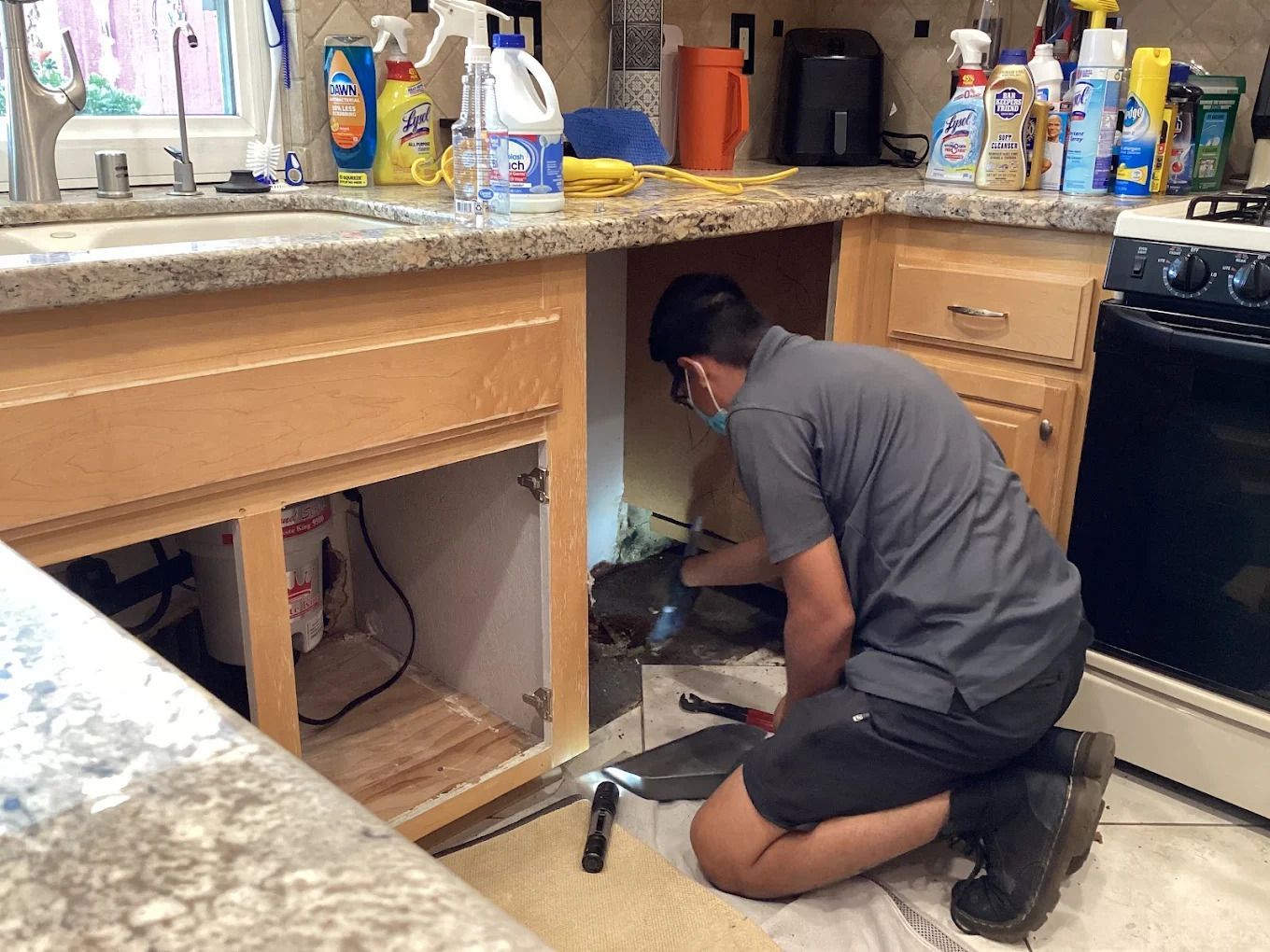Person kneeling under kitchen sink, repairing plumbing beside open cabinet and countertop clutter