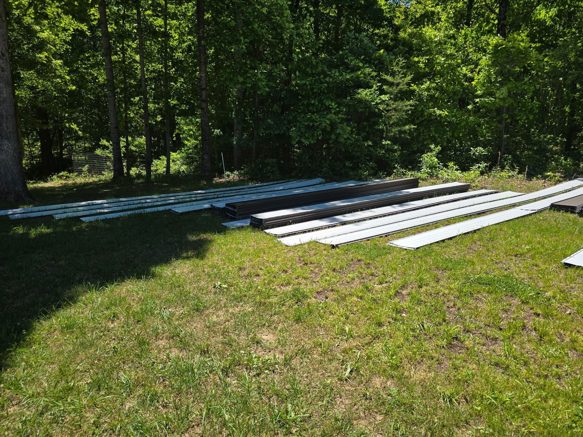 White metal sheets stacked on grass beside a wooded area in sunlight