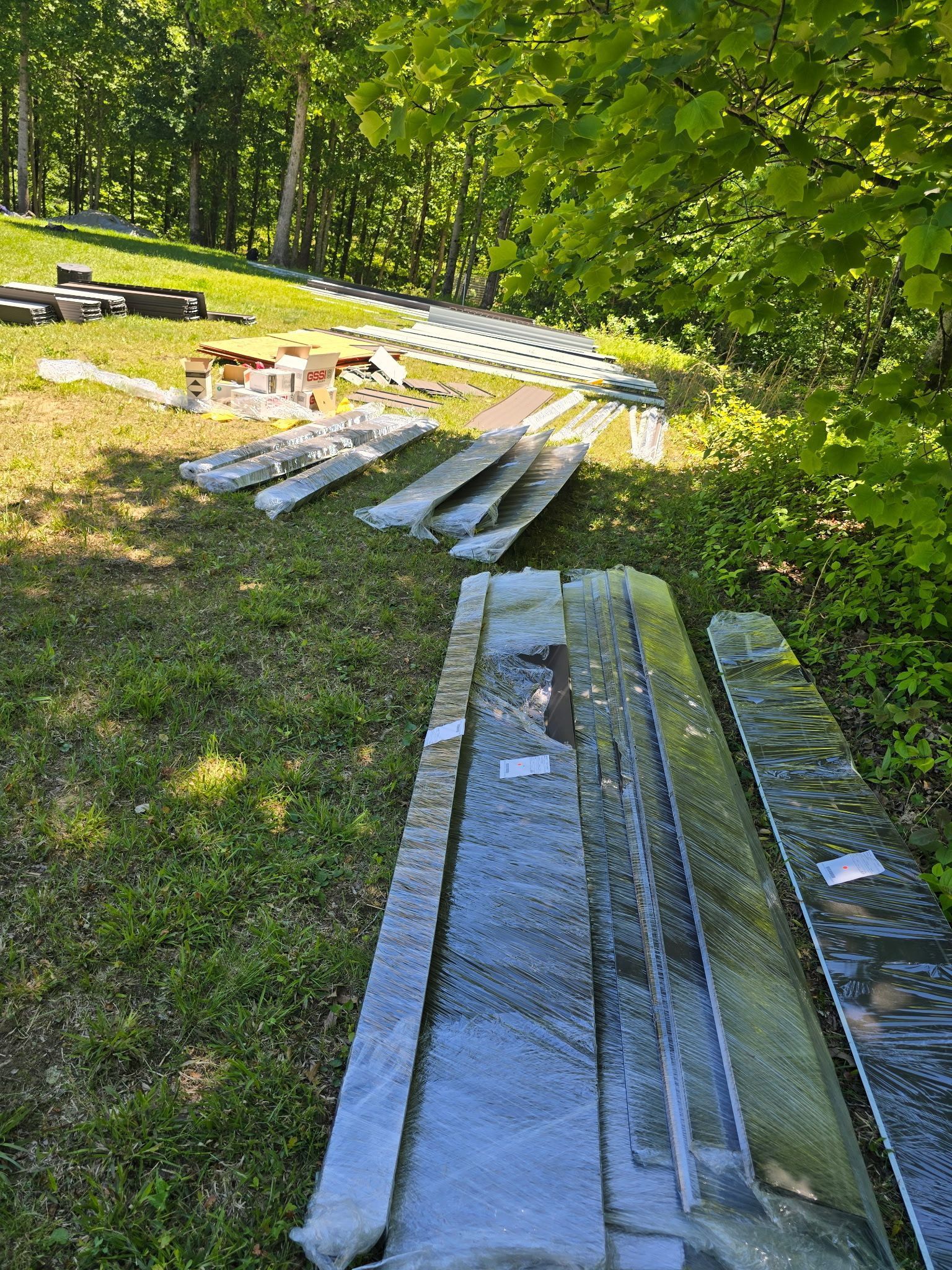 Stacked lumber and long glass panels on grass beside a sunlit wooded area.