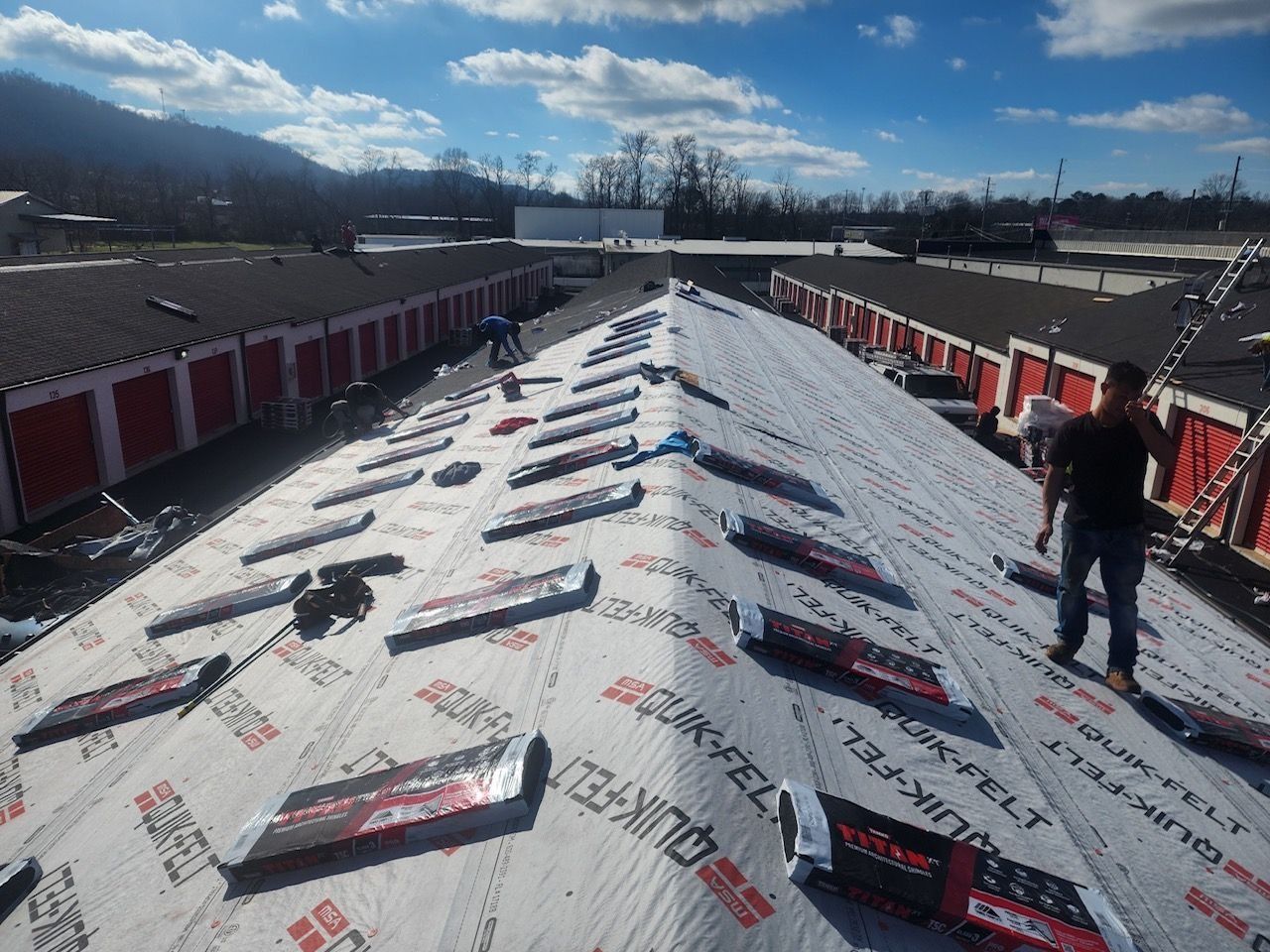 Flat commercial roof being repaired on a sunny day; white roofing material, tools, and equipment visible.