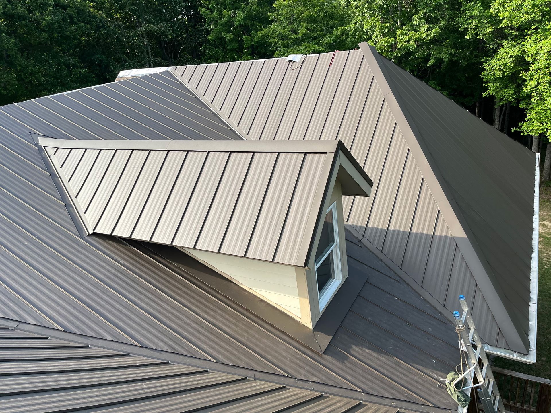 Gray metal roof with a dormer window and vent, surrounded by trees