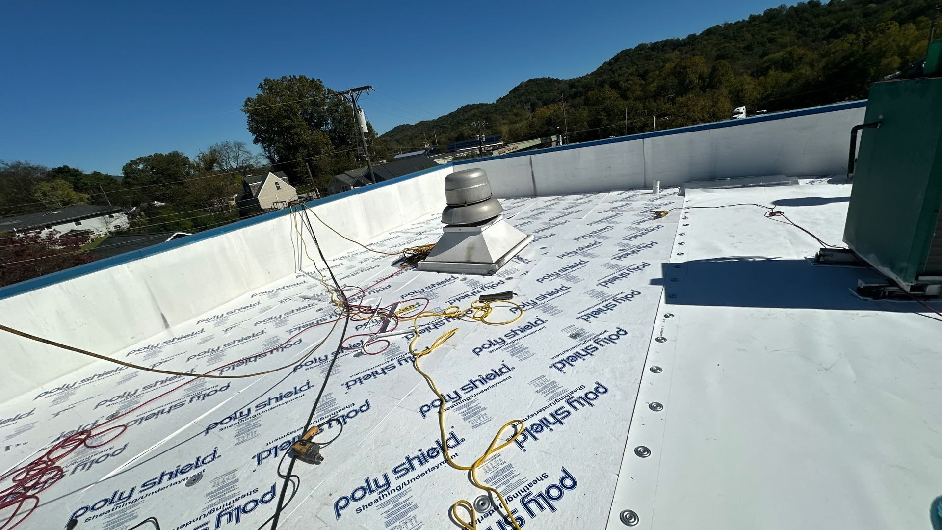 A rooftop with white protective covering, safety lines, and a vent, with a green backdrop of trees and a blue sky.