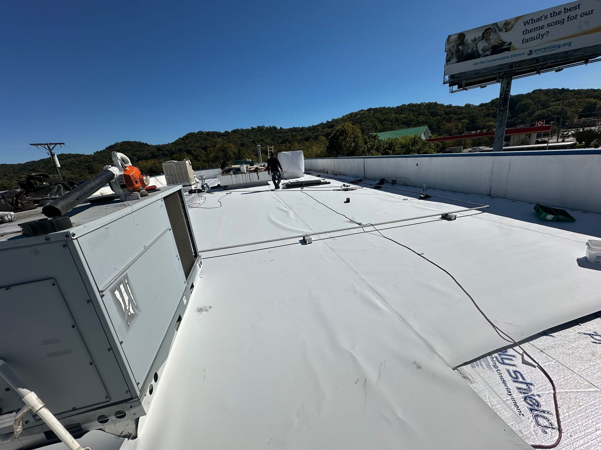 Workers on a white commercial roof on a sunny day near a billboard and trees.