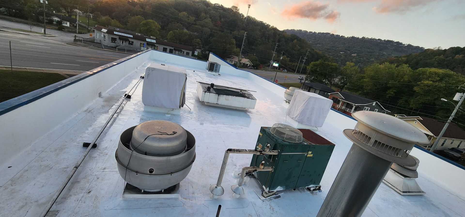 A white commercial rooftop with various mechanical units and equipment. The background features a road and wooded hillside.