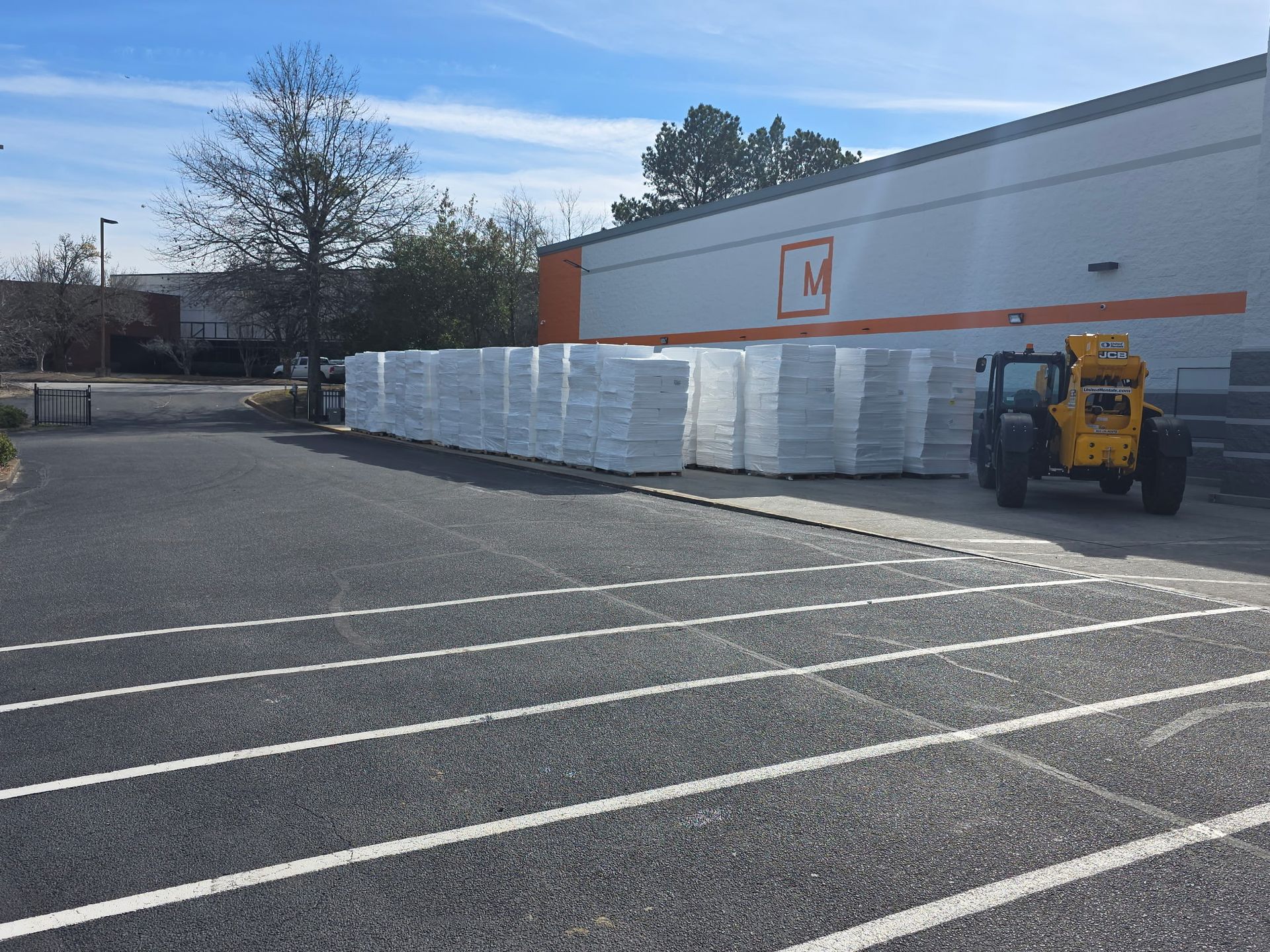 Piles of wrapped construction materials on pallets outside a building with an orange stripe. Sunlight.