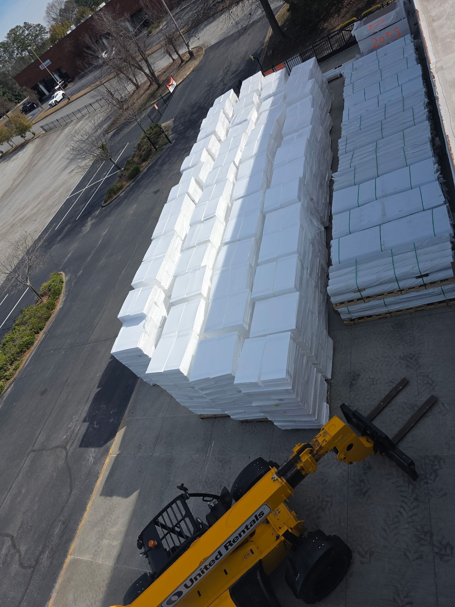 Exterior of a building with rolls of white material on pallets. A worker in a lift is near the wall.