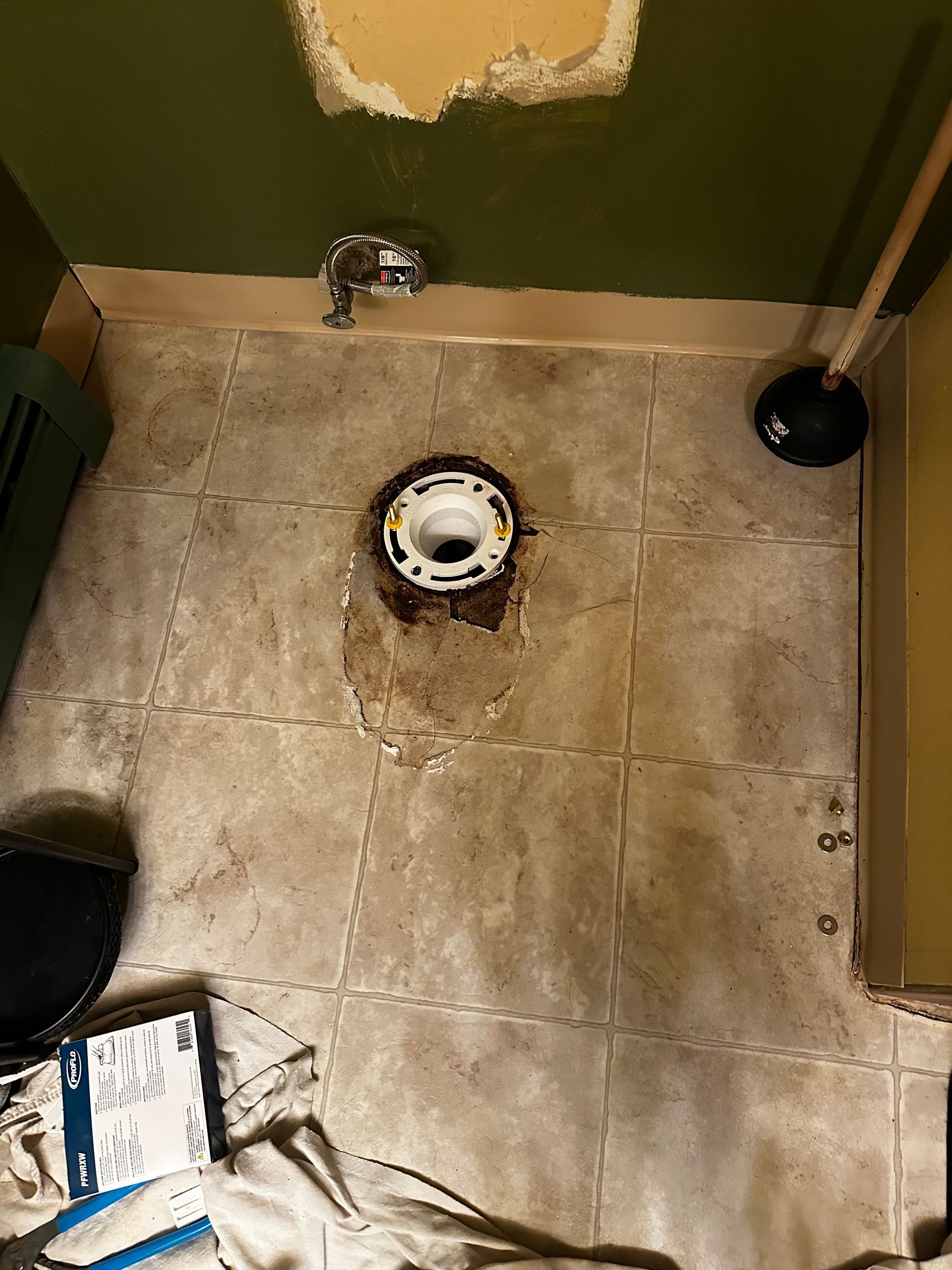 Toilet flange on bathroom tile with water stains, plunger, and repair materials visible.