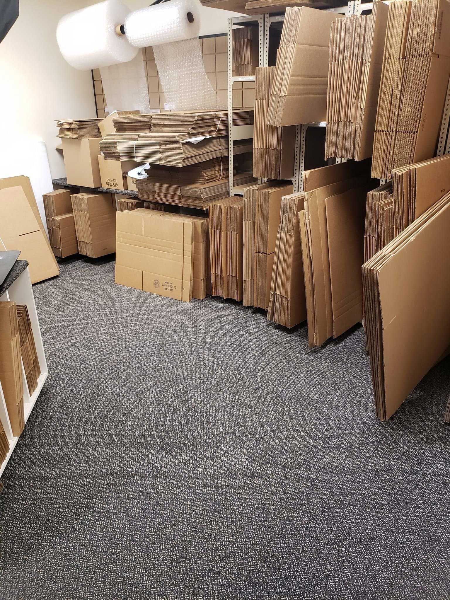 Cardboard boxes and packaging materials stored in a small room with grey carpet.