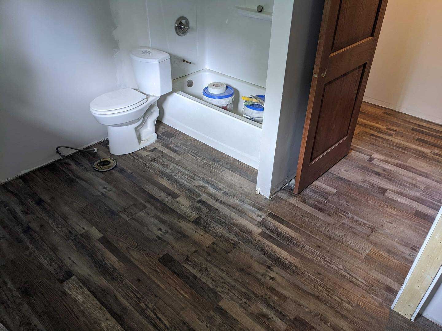 Bathroom with a toilet, bathtub, and wood-look flooring; ajar brown door.