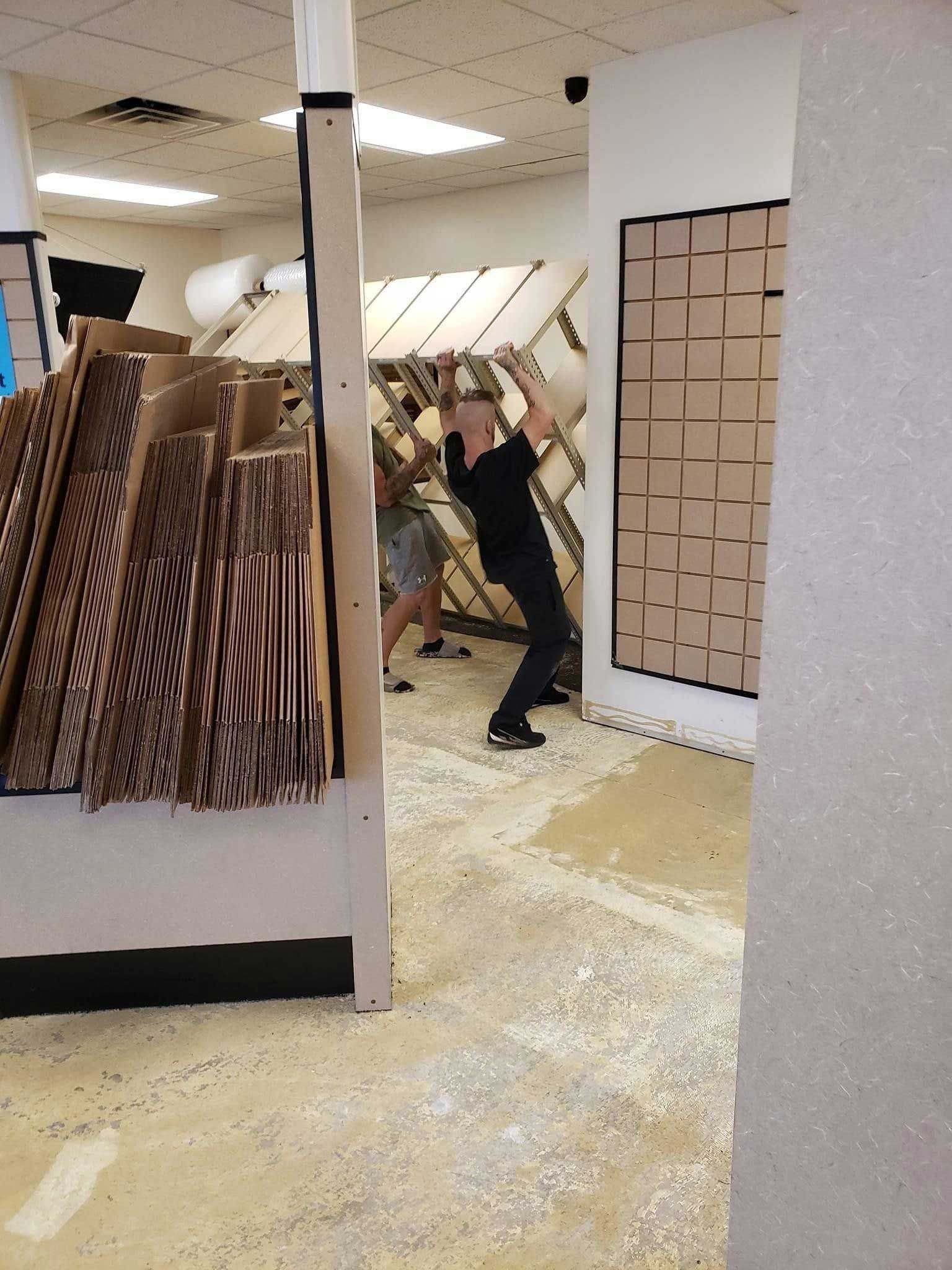 Man lifting heavy object in a messy room with wood debris on the floor; other person present.