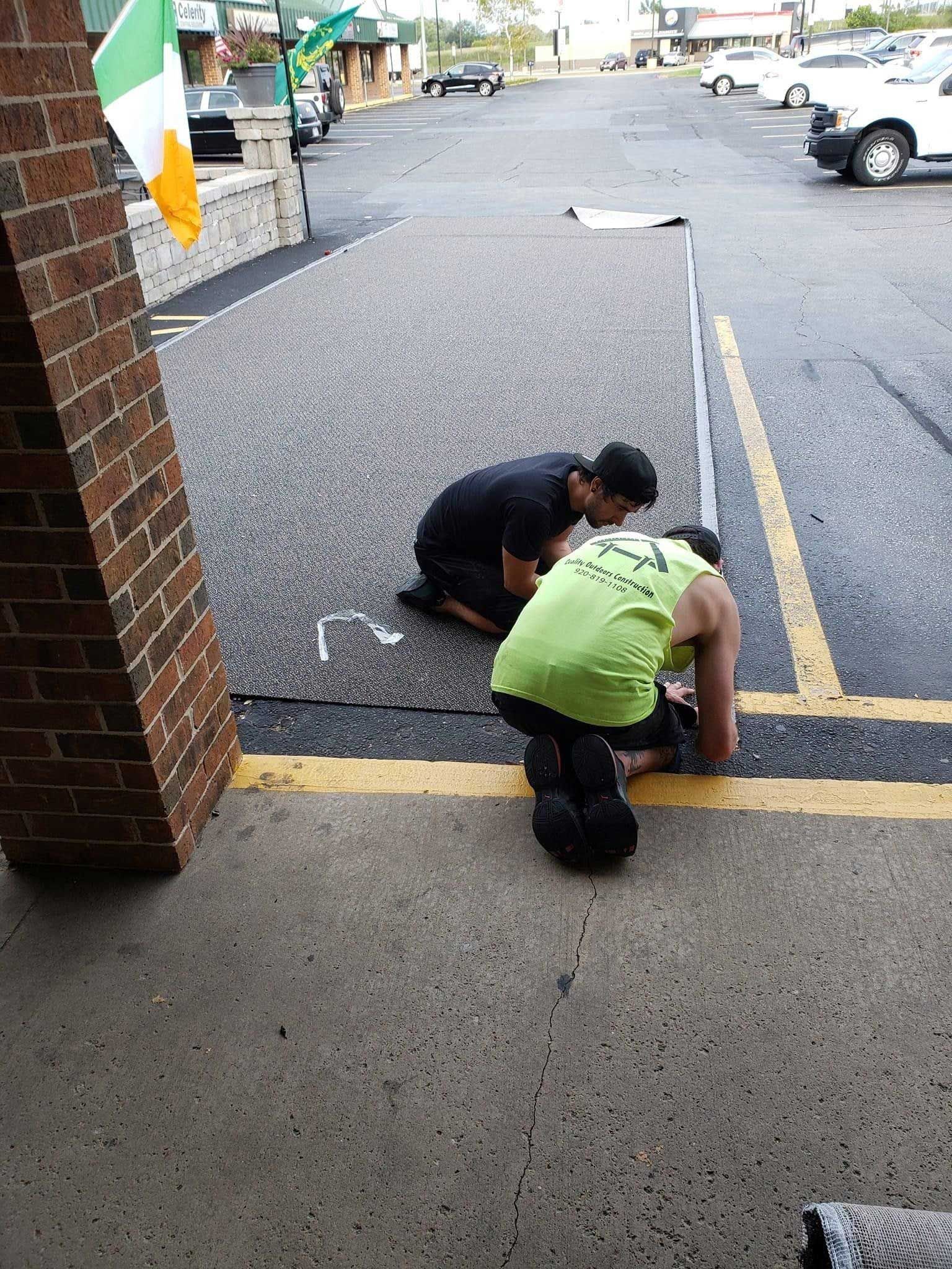 Two people kneel, working on a parking lot line. Green and black shirts, outside near building with Irish flag.