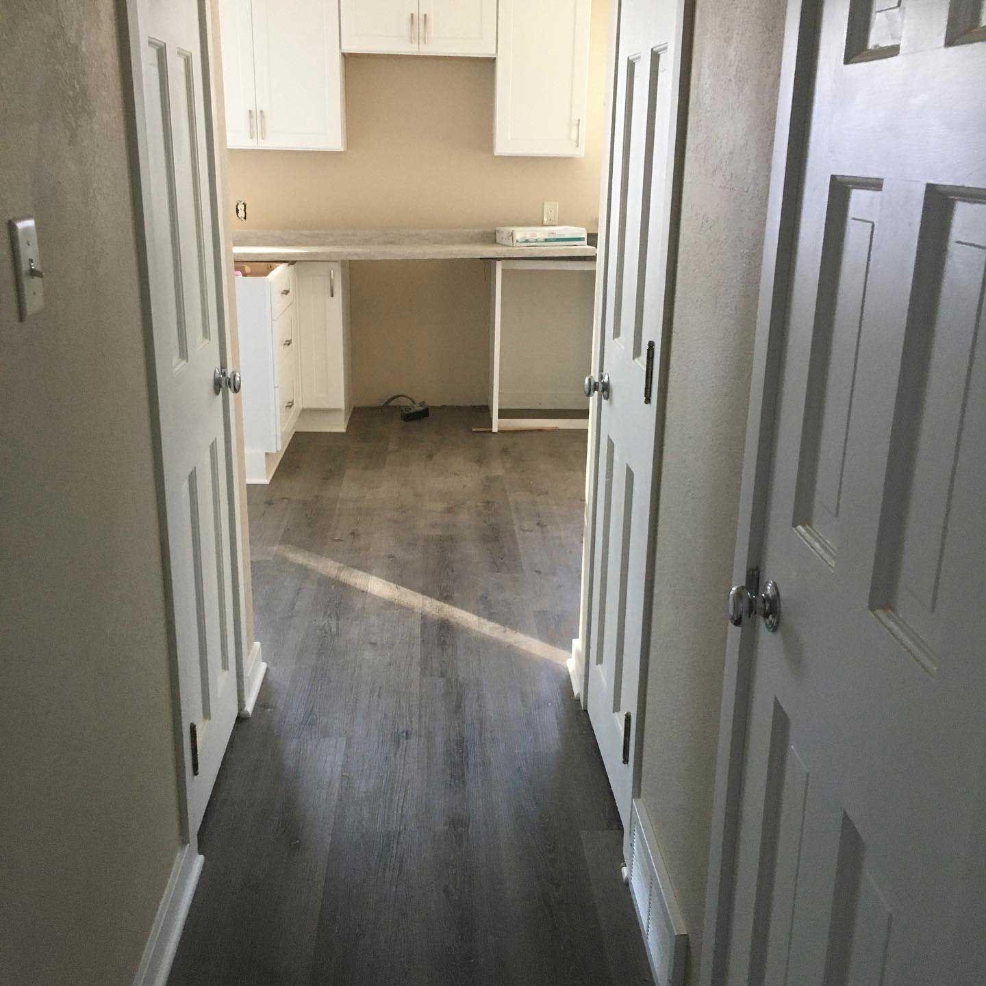 Hallway with gray wood flooring and white doors leading to a room with white cabinets and a workspace.