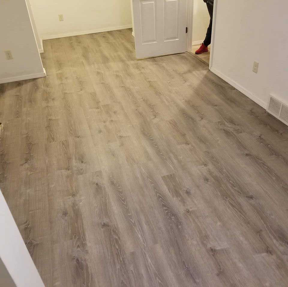 Light gray wood-look flooring installed in a room with white walls, a closed door, and a person's legs.