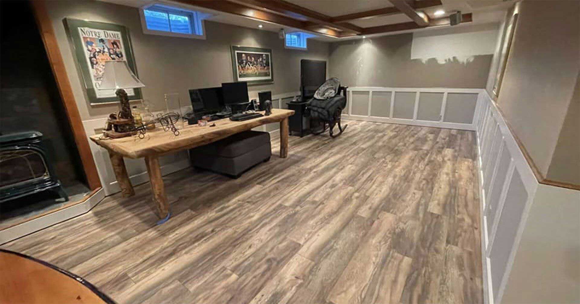 Finished basement with wood-look flooring, desk with computers, and decorative wall paneling.