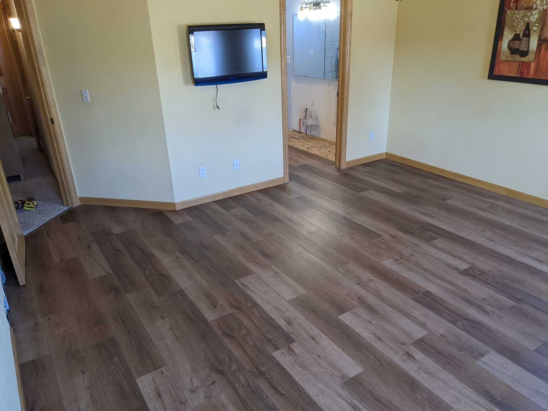 Empty room with brown wood-look flooring, beige walls, small mounted TV, doorway to a bathroom.
