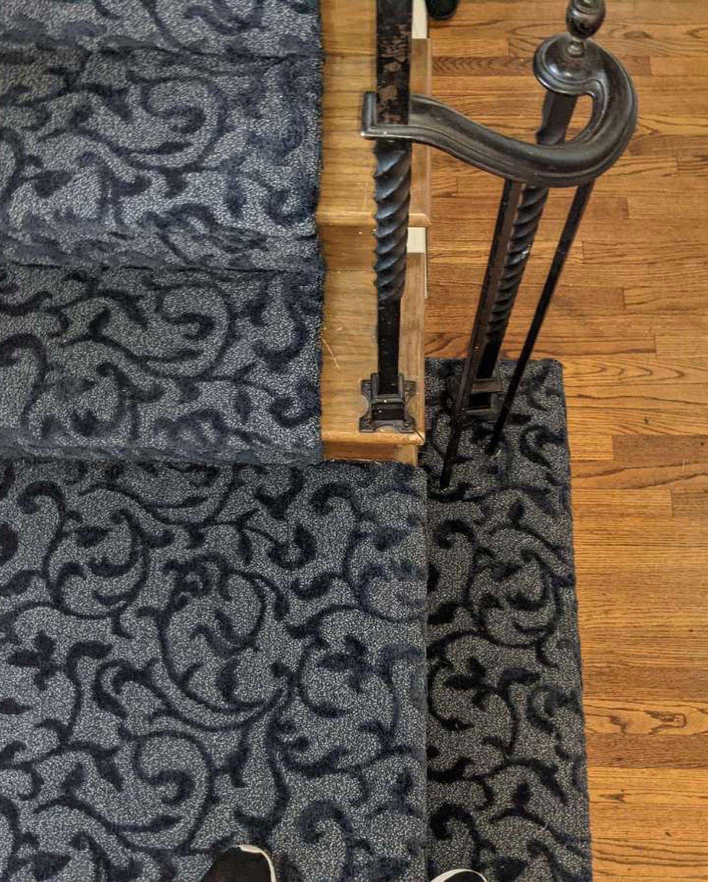 Carpeted staircase with floral pattern, dark grey and black, next to a wooden floor and metal railing.