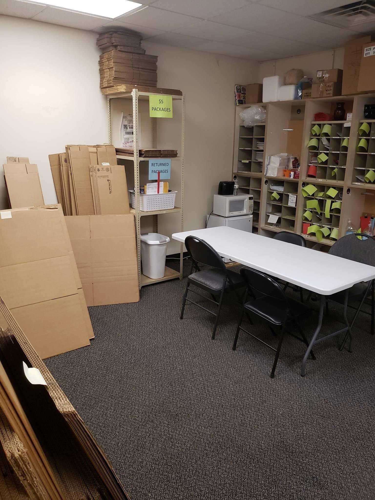 A small, cluttered office with cardboard boxes, shelves, a table, and chairs.