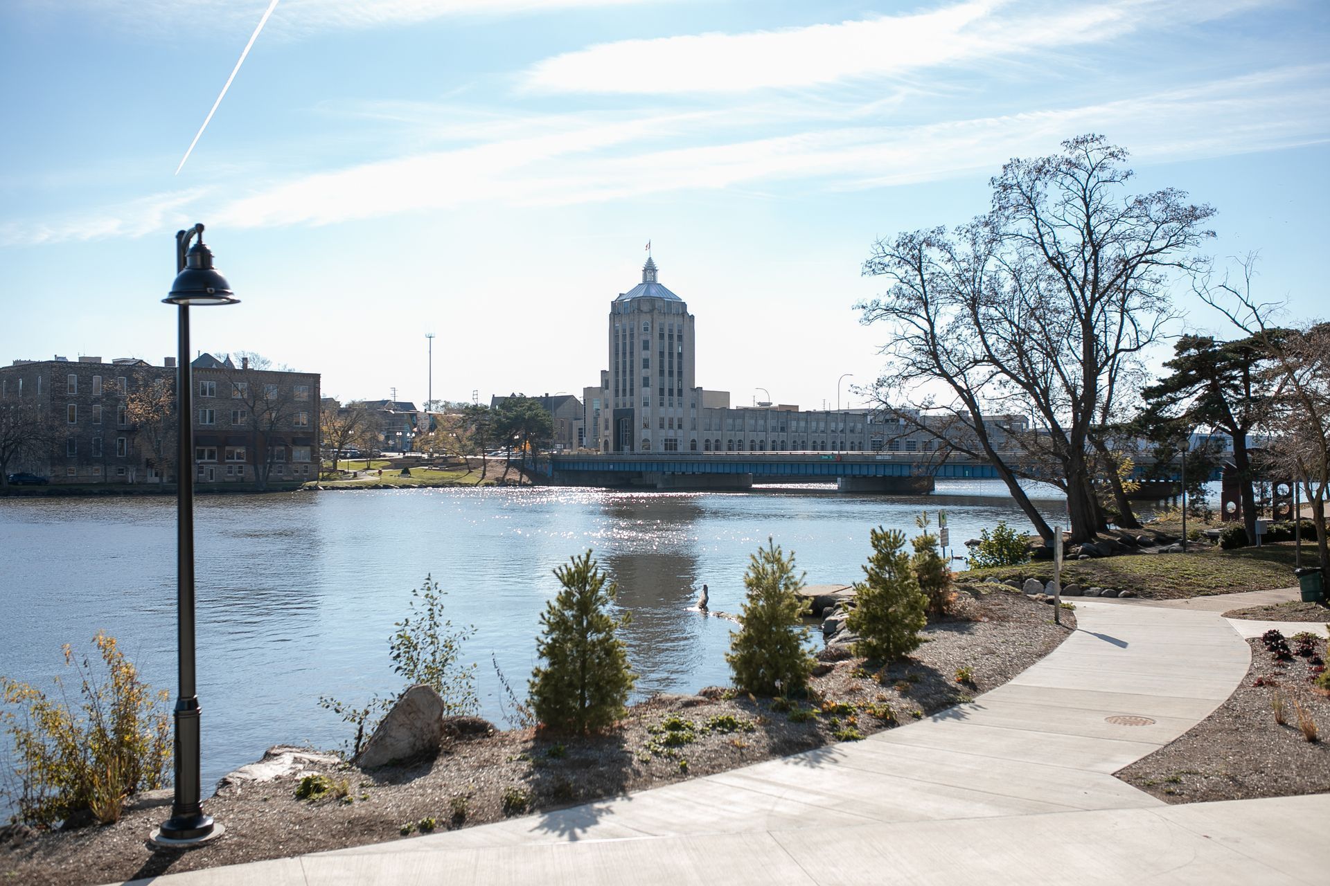 Park path overlooking water with a large building, trees, and a street lamp. Clear, sunny sky.