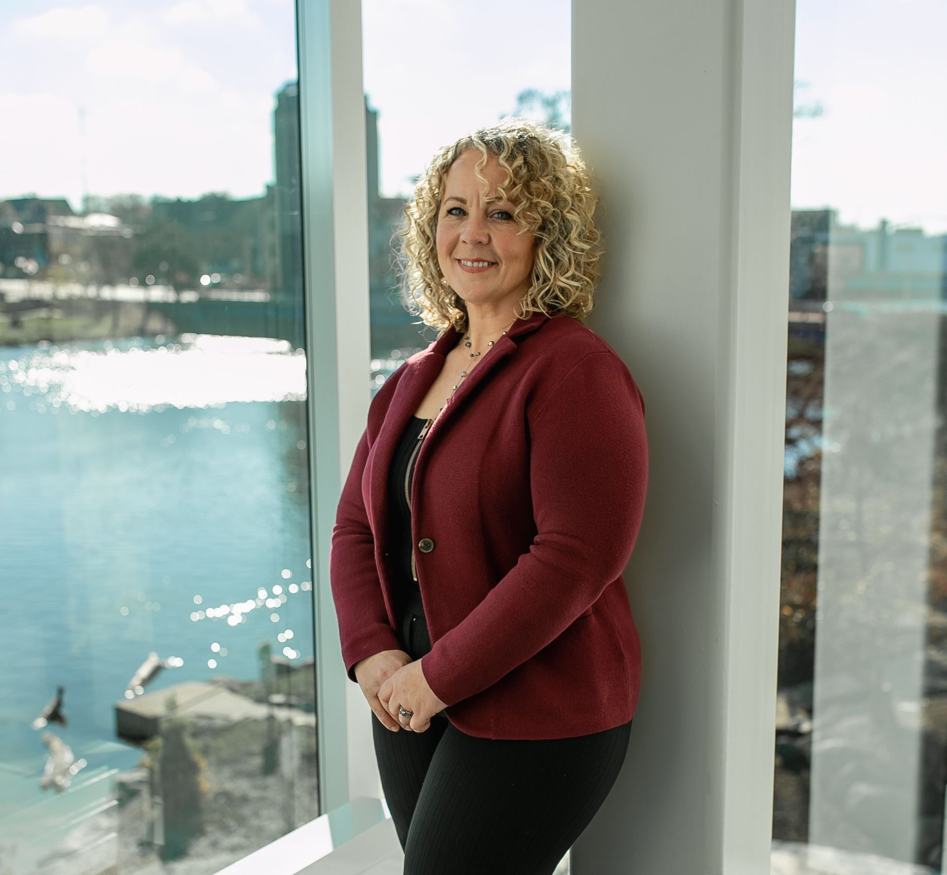 Woman with curly hair in maroon blazer leans by a window overlooking a river and city.