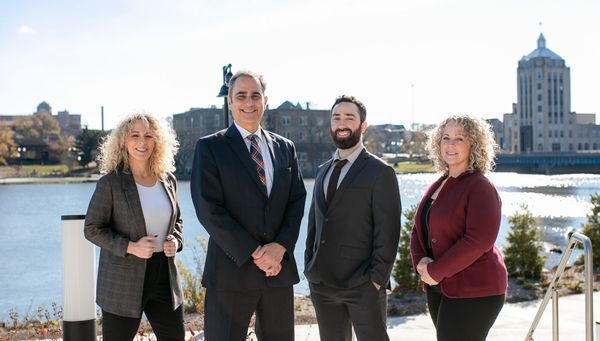 Four professionals posing outdoors by a river, with city buildings in the background.