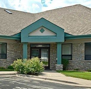 Front entrance of a stone building with teal trim and a glass door, framed by bushes and a paved walkway