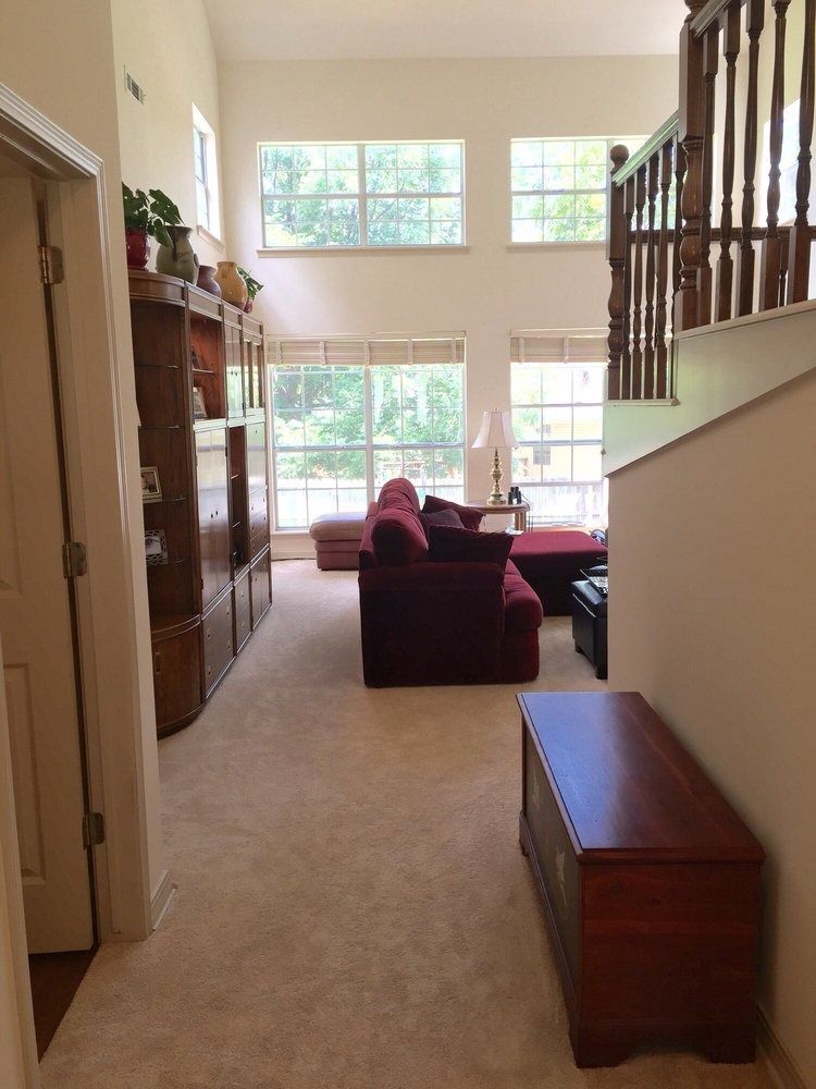 Bright carpeted foyer with staircase, red sofa, and large windows overlooking a living room.