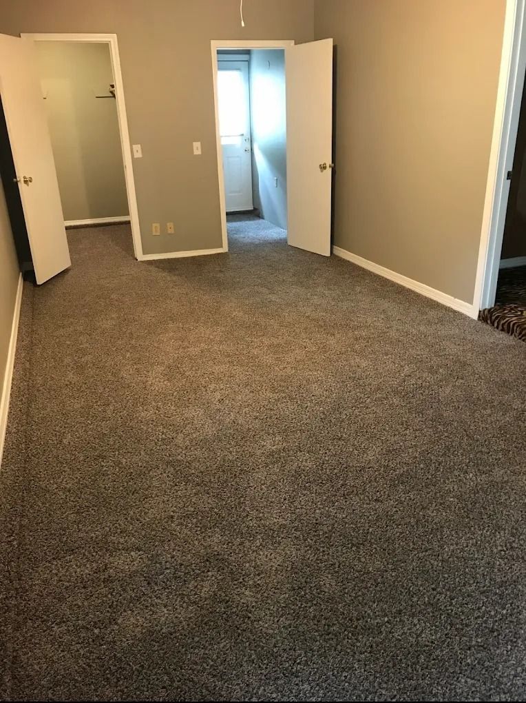 Empty beige room with brown carpet and open doors leading to a lit hallway and adjacent room