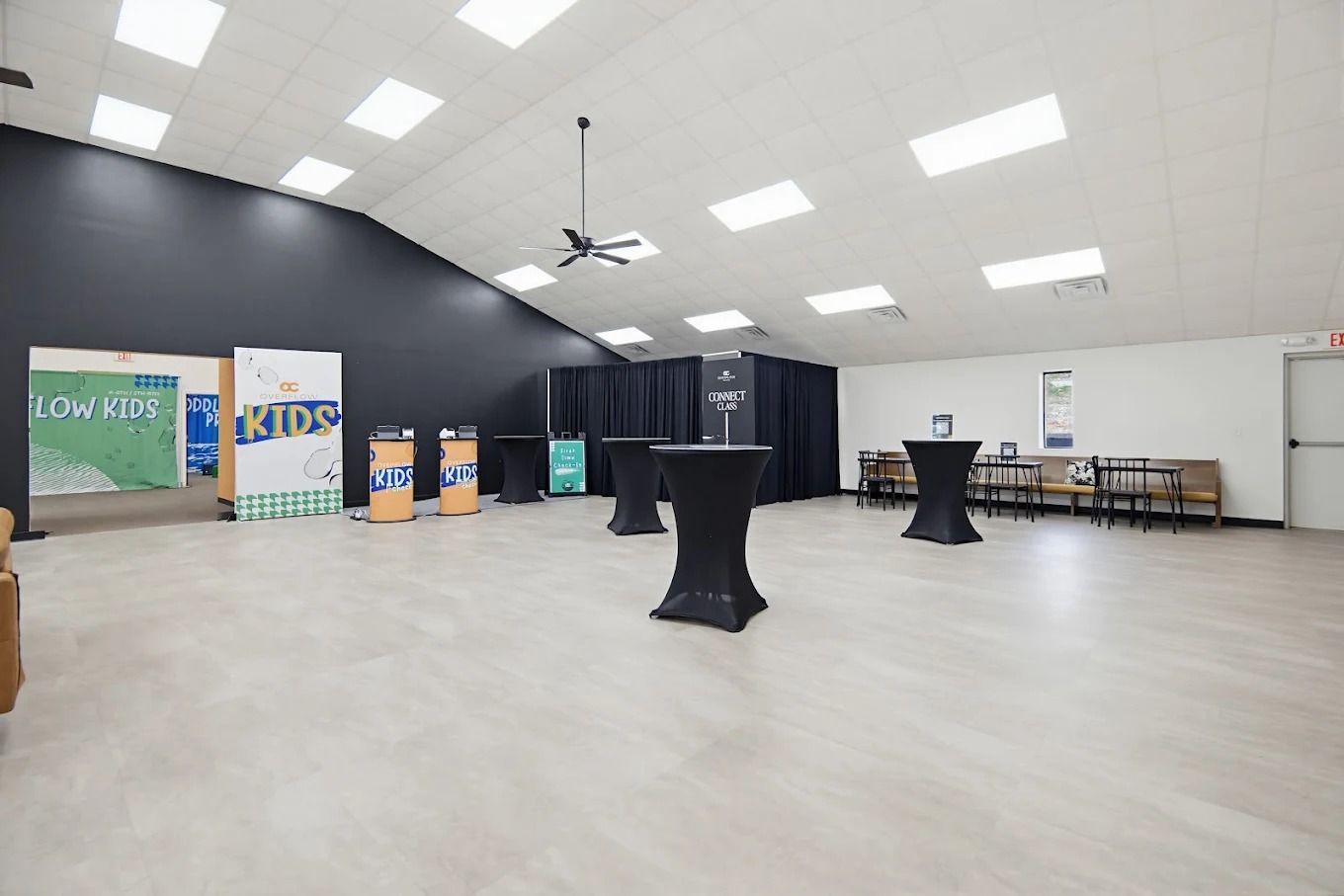 Empty event hall with high white ceiling, black drapes, tables, and a small stage area with banners and signs