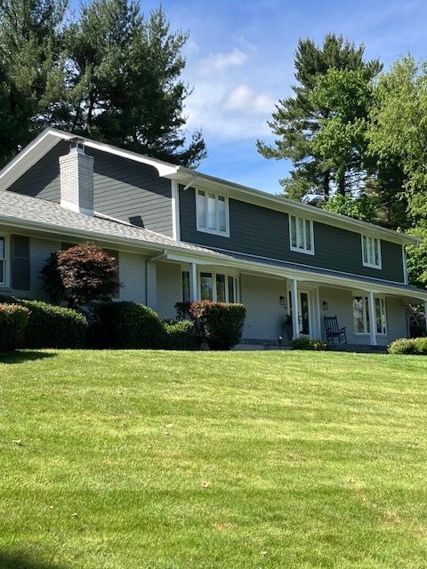 Two-story house with green siding and a gray roof, set on a green lawn with trees in the background.