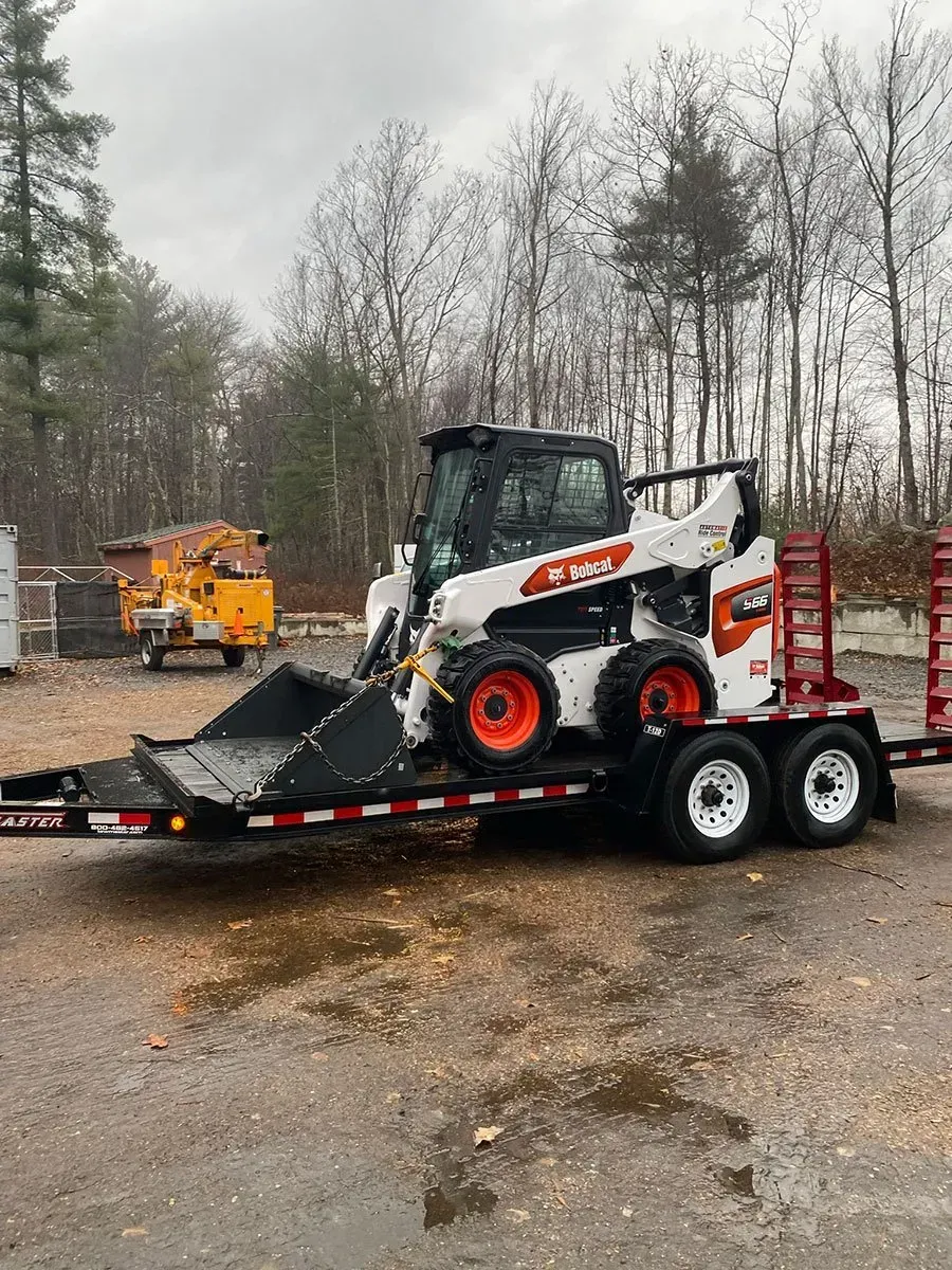 Bobcat skid-steer loader on a trailer in an outdoor area; orange wheels, overcast sky.