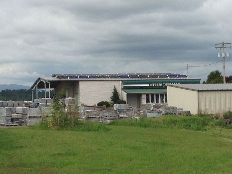 Building with solar panels on the roof, exterior view. Gray sky, grassy field, and stacks of materials in the foreground.