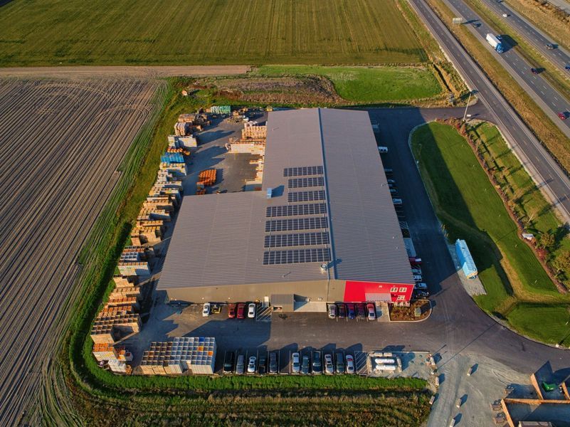 Aerial view of a large commercial building with solar panels, surrounded by a parking lot, fields, and a highway.