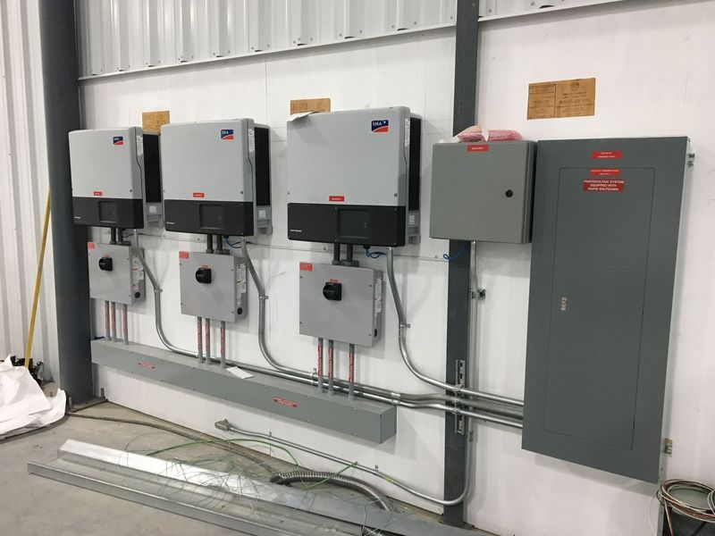 Solar inverters, disconnects, and electrical panel mounted on a white wall in a building.