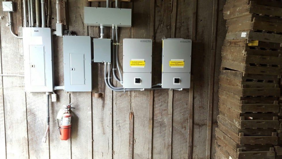Electrical equipment, including panels and inverters, mounted on a weathered wooden wall. A fire extinguisher is nearby.